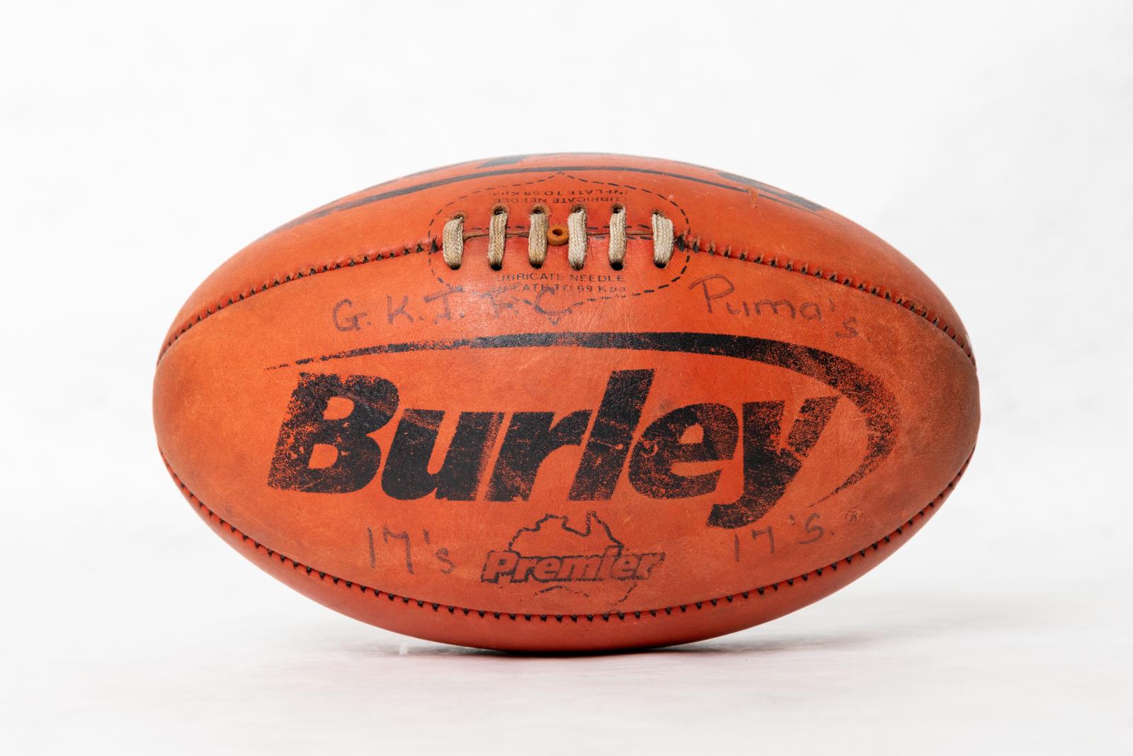 Red Australian Rules 'Burley' football. Black text is worn, and states 'Burley', with 'Premier' on top of an Australian map outline, the top of the football states ' LUBRICATE NEEDLE/ INFLATE TO 69 Kpa, duplicated on either side of the stitching. 