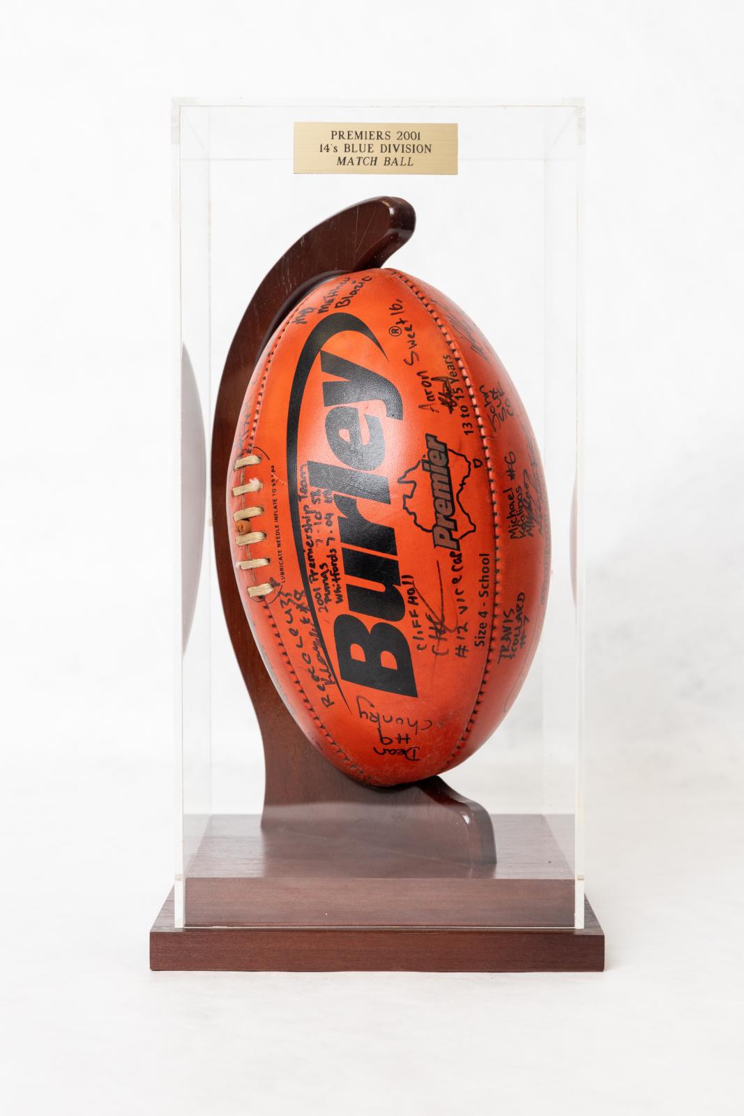 A red 'Burley' football held upright in a curved wooden stand, encased within a clear Perspex box. There is a small metal plate affixed to the front of the Perspex box near the top, which states 'PREMIERS 2001/ 14'S BLUE DIVISION/ MATCH BALL'. The football is signed in black marker by the players and coaches.