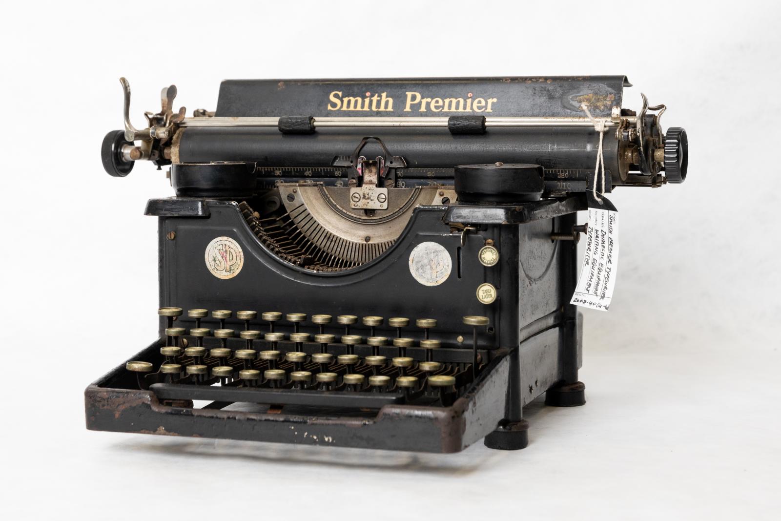 Smith Premier manual typewriter. Painted black metal with beige coloured plastic coated keys. Two metal spools for holding black and red printer ribbon, long round rubber roller and black plastic knob for holding paper. On the front are two metal discs with 'SP' in centre. 'Smith Premier' printed in gold text on the top metal flap that covers silver metal mechanisms at the back. 