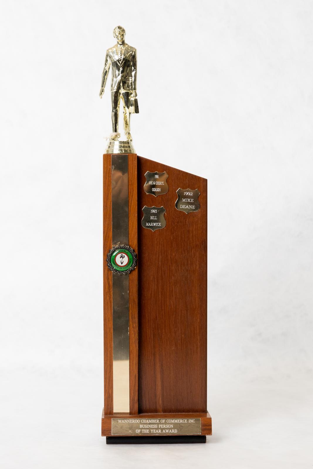 A perpendicular Jarrah timber trophy with gold metal shields, long metal strip and award details attached with glue to trophy. A plastic gold coloured figurine of a businessman sits on top of the trophy. 