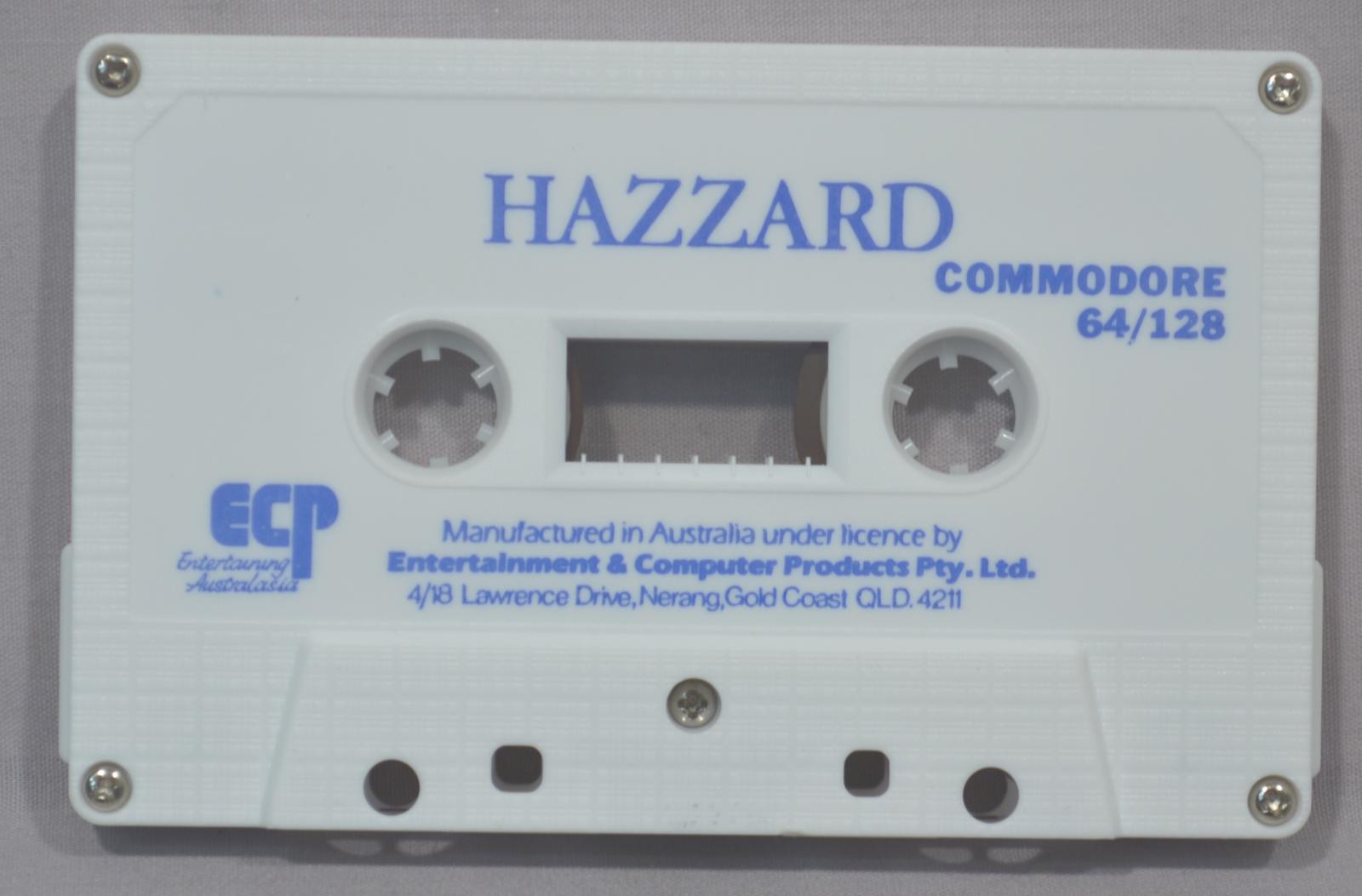 Rectangular white plastic cassette tape. Label on top with game name (Hazzard) and manufacturer details on the bottom.