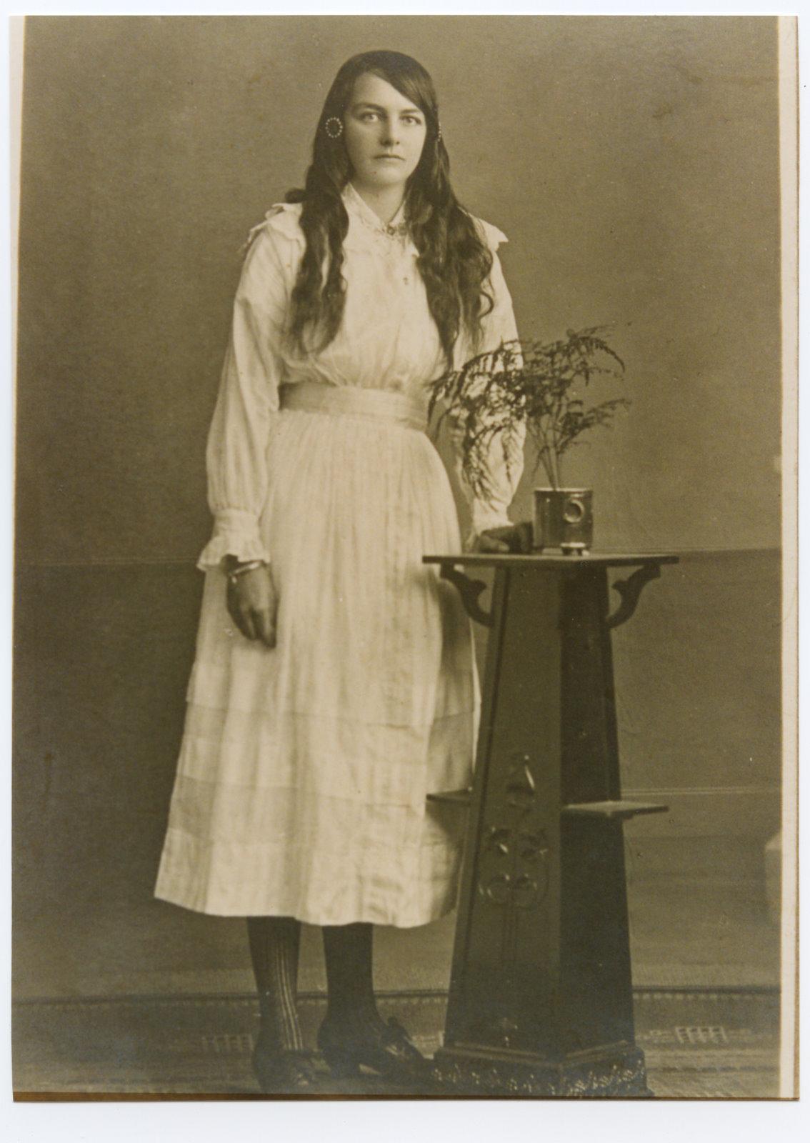 Black and white studio photograph of Teresa May Cosgrave age 17 wearing a white long sleeve dress, belted at the waist. She has long dark hair that is draped over each shoulder. She is standing next to a wooden plynth with a potted plant in it. 