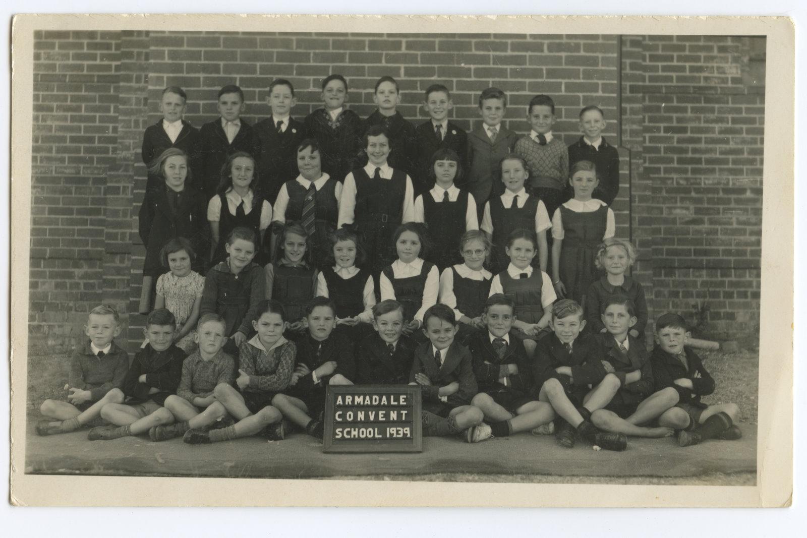 Black and white photograph of four rows of children, boys and girls, in front of a brick wall. Sign in front of the front row says Armadale Convent School 1939