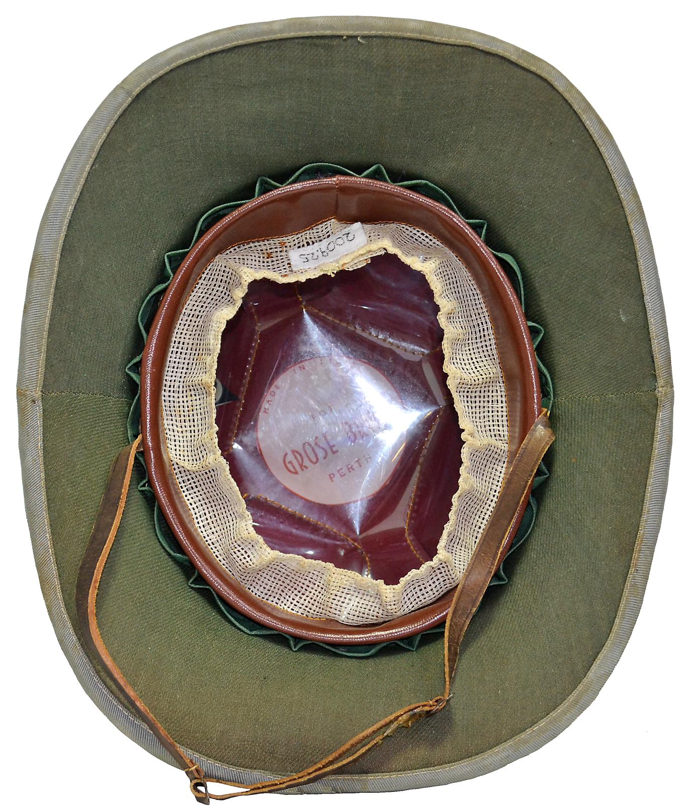 Inside of an oval shaped pith helmet. Green cloth covers the underside of the brim, a brown leather inlay is on the inside of the helmet, while a band of white cloth mesh is around the inside the the helmet brim. A brown leather chin strap is attached to the two sides of the inner brim