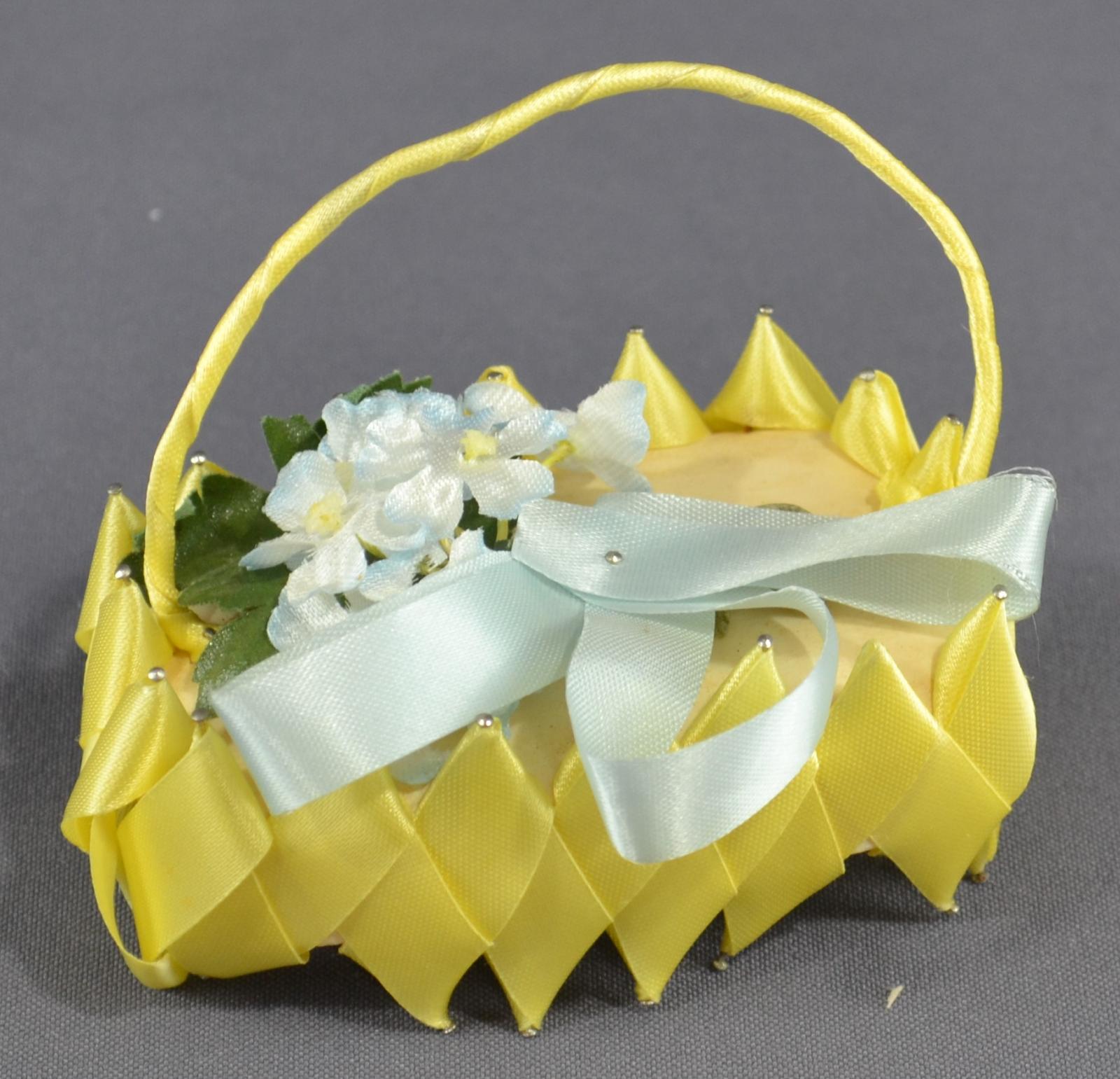 Cake of soap with pins inserted to upper and lower edge, around them is laced yellow ribbon in a criss cross pattern. A handle of the same yellow ribbon is pinned to the ends. Pale blue flowers with green foliage and pale blue ribbon is pinned to the upper surface