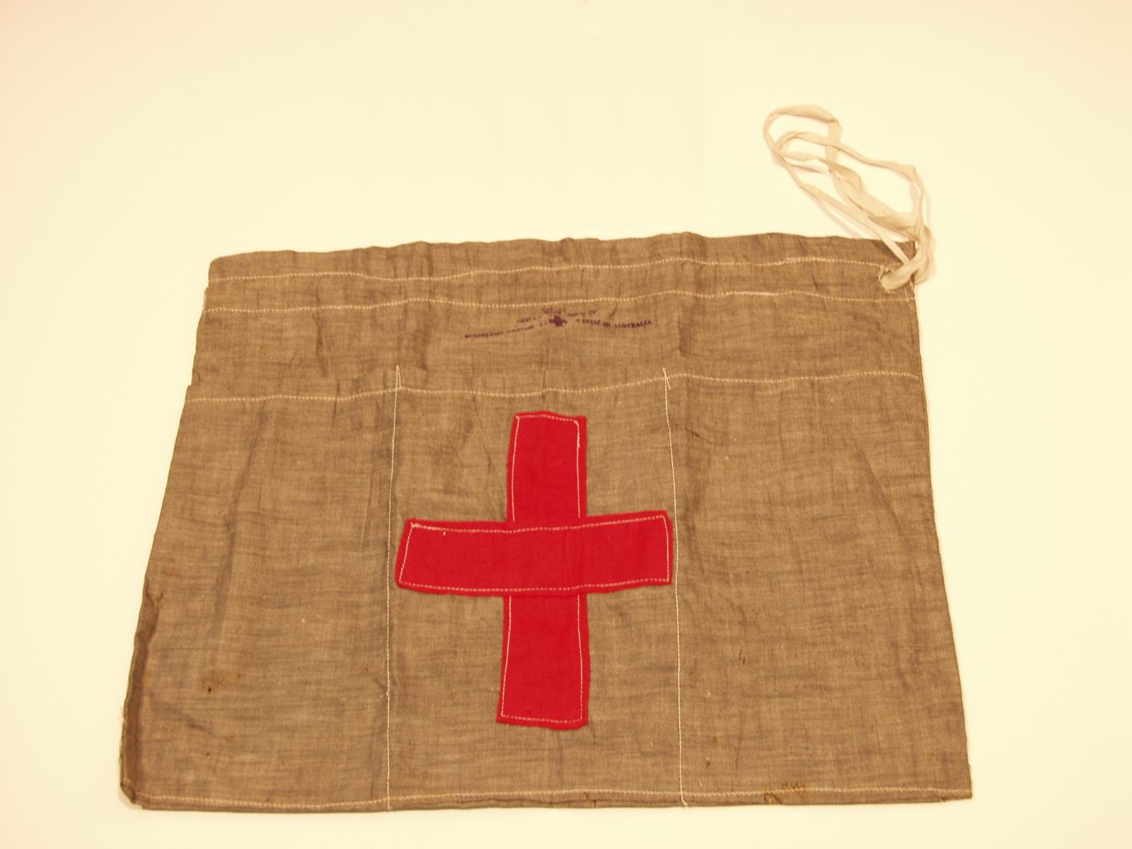 Light denim coloured rectangle cloth bag with a red fabric cross in the middle and a white draw string on the right side of the opening at the top