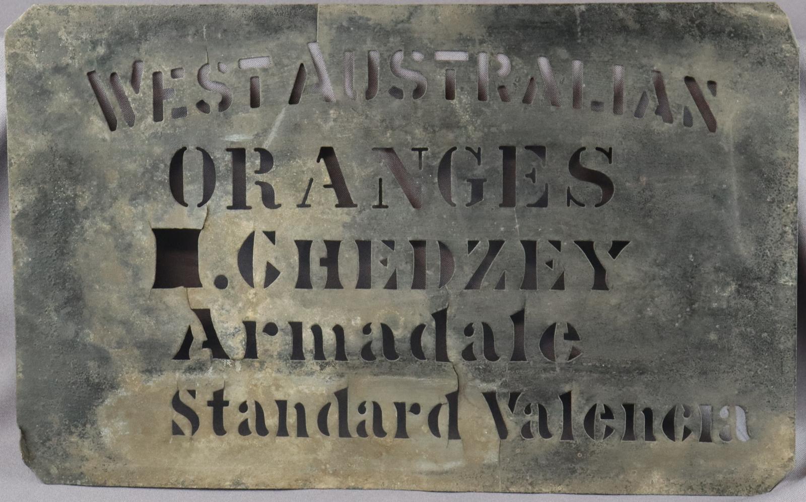 Flat rectangle sheet of metal with five lines of text cut out to for a stencil. Text says  [WEST AUSTRALIAN ORANGES CHEDZEY Armadale Standard Valencia ]