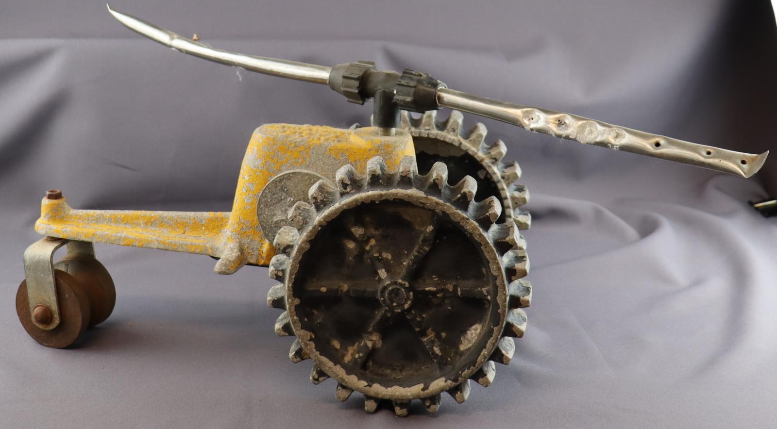 Yellow bodied tractor sprinkler. On top are two metal sprayer arms, at back two black tires, at front a single metal roller wheel.