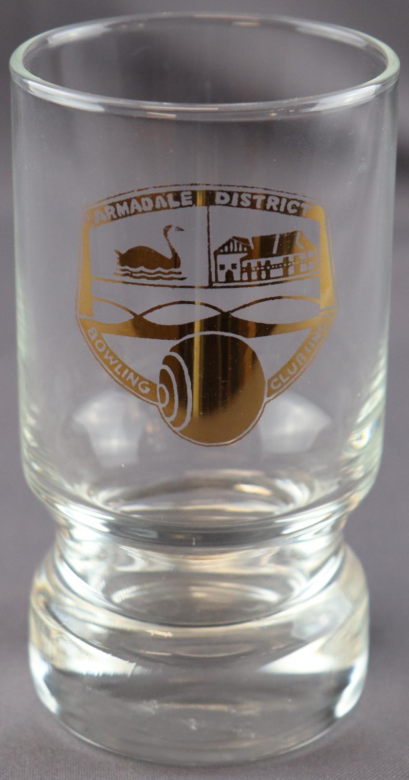 Clear glass  cylindrical shot glass with Shield shaped logo on front.Logo divided into three parts, top left features a swan, top right features illustration of the Narrogin Inn, Bottom part features illustration of a bowl