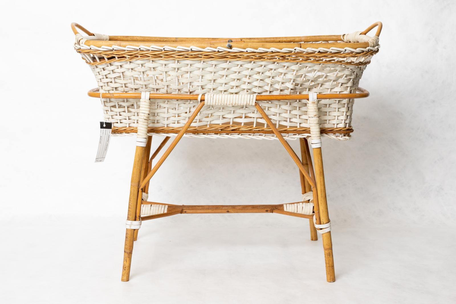 A doll's cradle on a stand, all made from rattan and white plastic, the white plastic is weaved to make the body of the cradle and to hold together the wood.