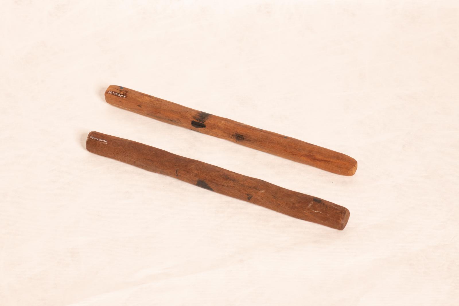 Pair of dark wood clapping sticks, thin elongated shape with rounded sides and painted linear pattern near centre.