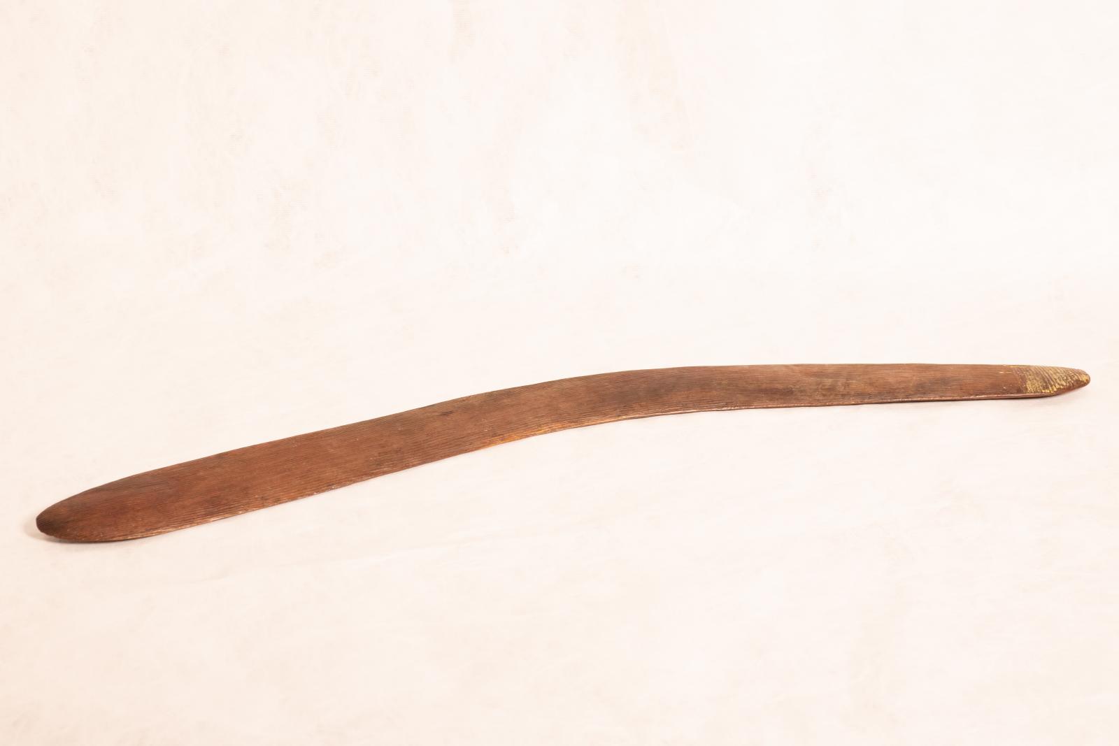 Carved wooden boomerang with rounded edges and ends. Raised linear pattern running from end to end along length of boomerang.