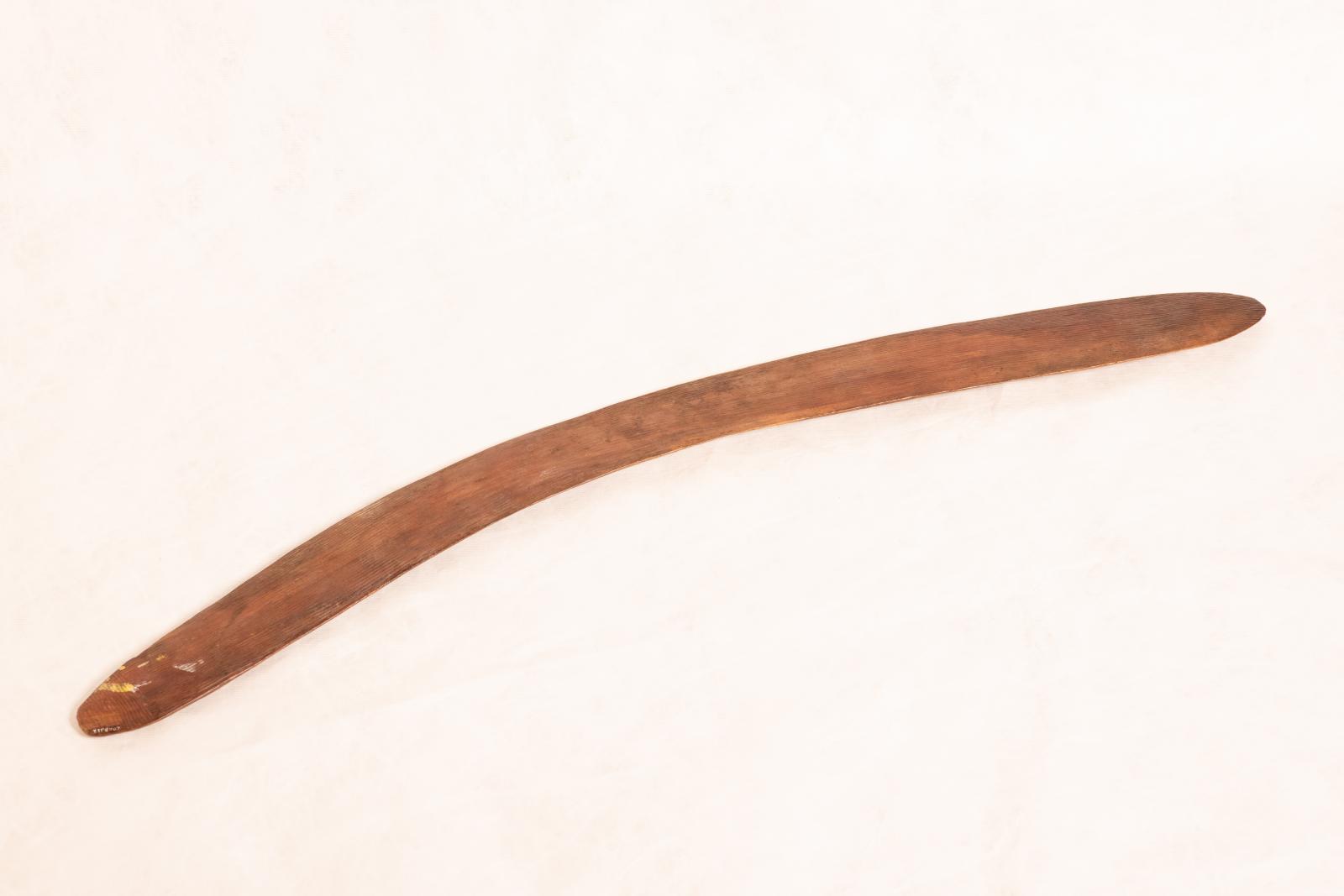 Carved wooden boomerang with rounded edges and ends. Raised linear pattern running from end to end along length of boomerang.