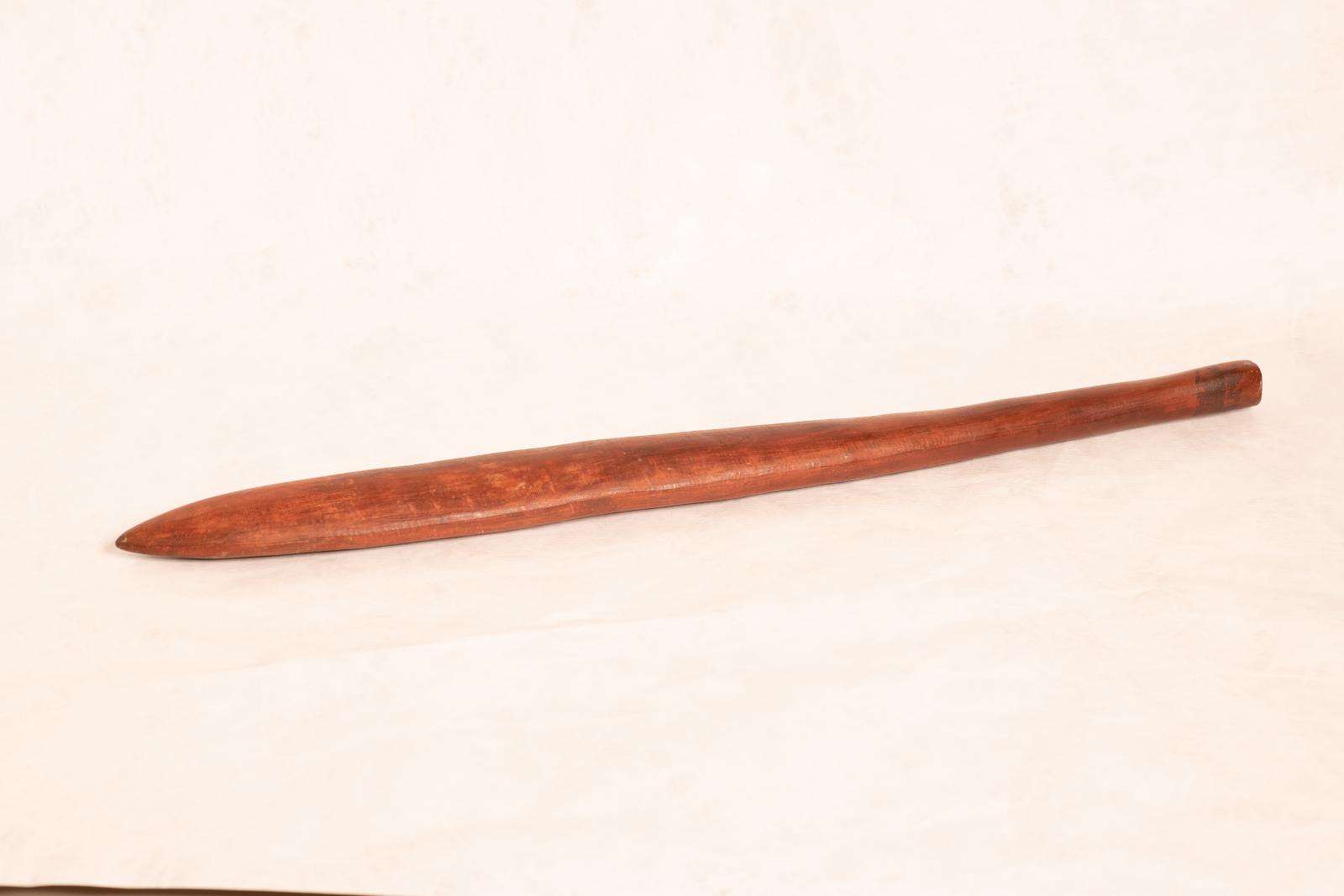 Wanna, a carved wooden digging stick. The flat pointed end tapers to a round handle and there are small linear scratchings on the surface of the wood.