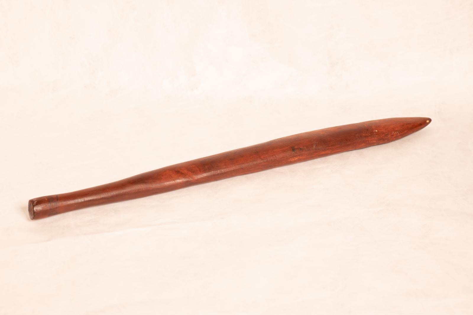 Wanna, a carved wooden digging stick. The flat pointed end tapers to a round handle and there are small linear scratchings on the surface of the wood.