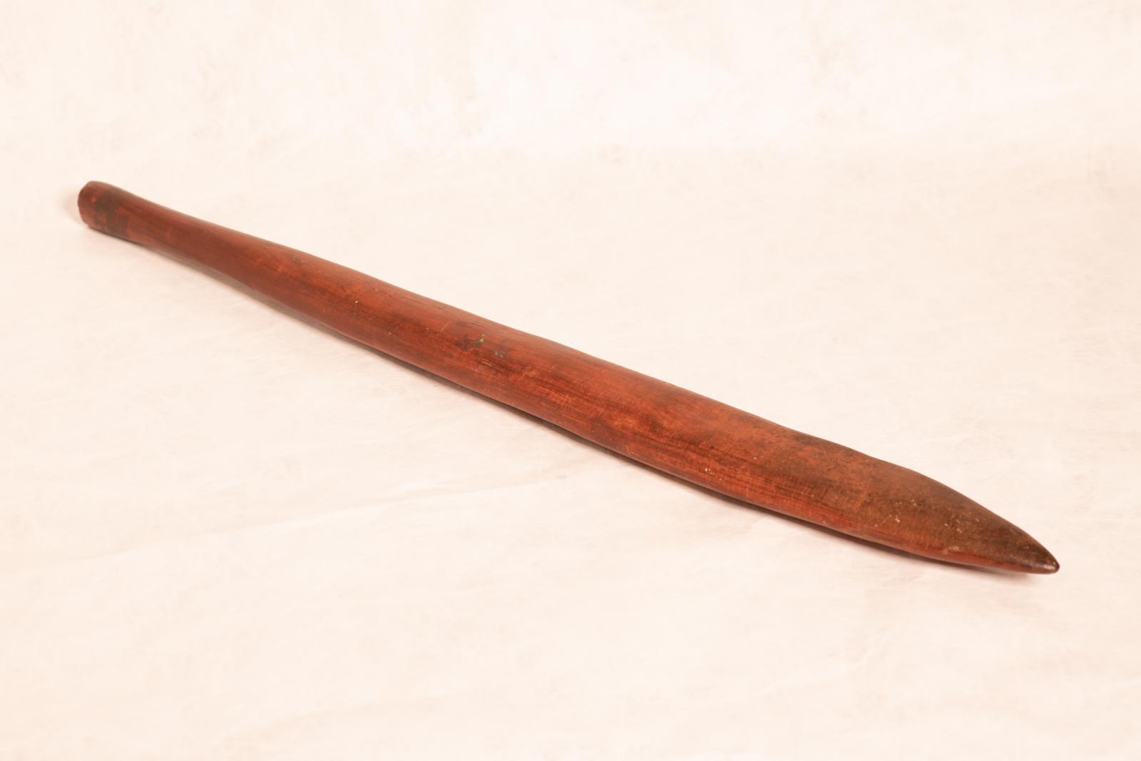 Wanna, a carved wooden digging stick. The flat pointed end tapers to a round handle and there are small linear scratchings on the surface of the wood.