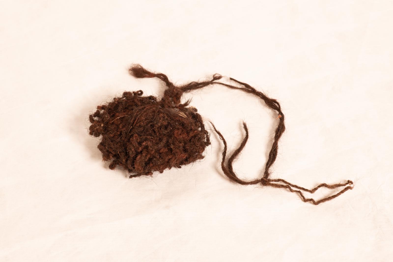 Woven, brown human hair pubic cover wound into a large bunch with long twisted strands of hair coming from each side. Small white feathers are woven through twisted strands.