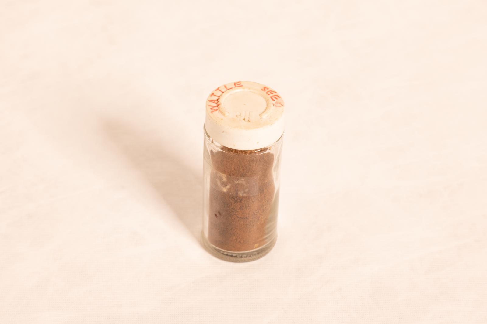 Clear glass jar with white plastic lid and dried wattle seeds inside. 'WATTLE SEED' written on the top of the lid in orange texta