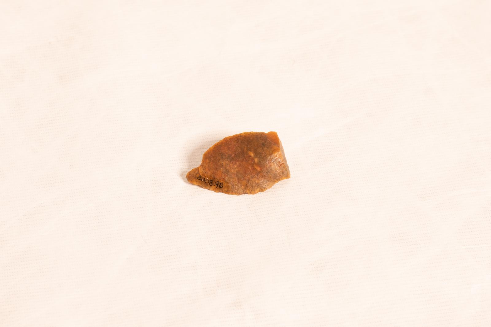 Small carved beige stone spearhead with two long sides tapering to a point and short square end. Sides are carved to a sharp edge. The point has a small chip from it on one side.