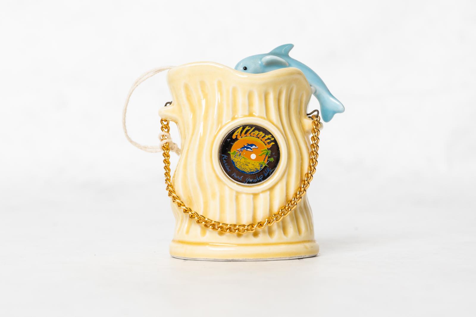 A cream coloured ceramic ornament with a fluted pattern. The item is open at the top and there is a blue ceramic dolphin resting on the right hand side lip facing the inside of the ornament. There are two small handles on either side, the left is higher than the right and connected just above these handles is a metal clip inserted into a hole. There is a gold chain connected to each metal clip which can be used as a handle. On the front side is a circular medallion with words 'Atlantis/Marine Park, Yanchep 