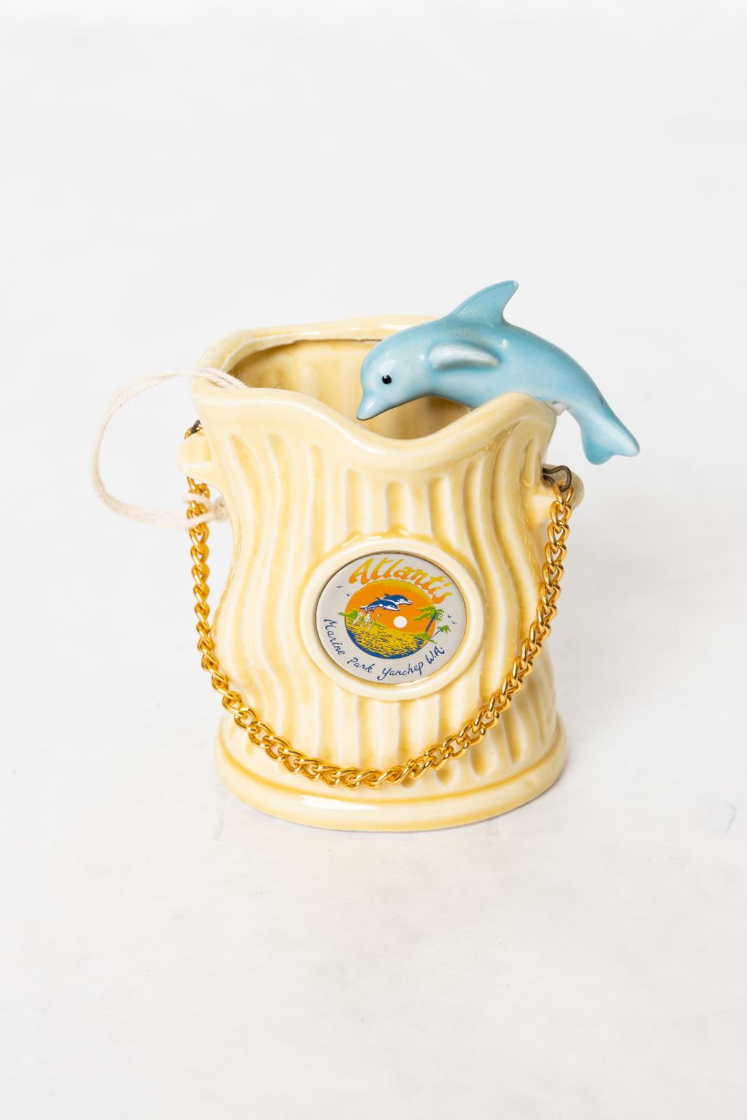 A cream coloured ceramic ornament with a fluted pattern. The item is open at the top and there is a blue ceramic dolphin resting on the right hand side lip facing the inside of the ornament. There are two small handles on either side, the left is higher than the right and connected just above these handles is a metal clip inserted into a hole. There is a gold chain connected to each metal clip which can be used as a handle. On the front side is a circular medallion with words 'Atlantis/Marine Park, Yanchep 