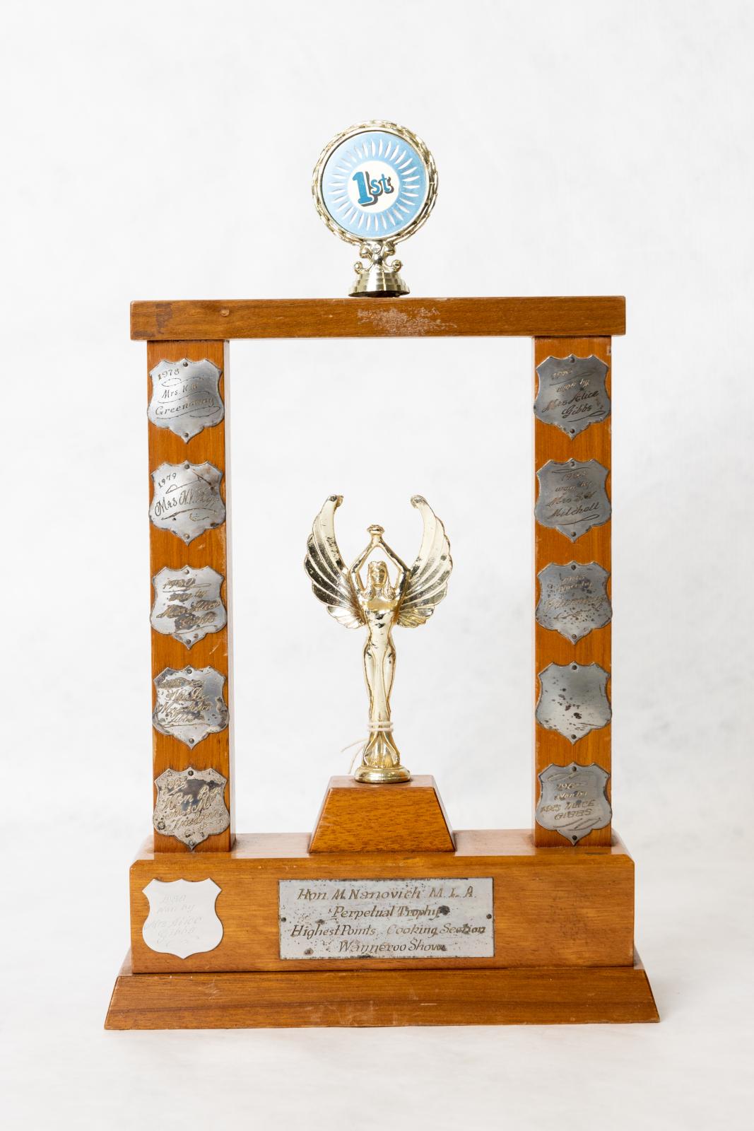 A rectangular shaped trophy, with a column on the left hand and right hand sides with a cross bar on the top. The top cross bar has a small circular medal with 1st written in blue in the centre. Attached to the base, between the two upright wooden posts sits a metal 'angel' styled figurine.