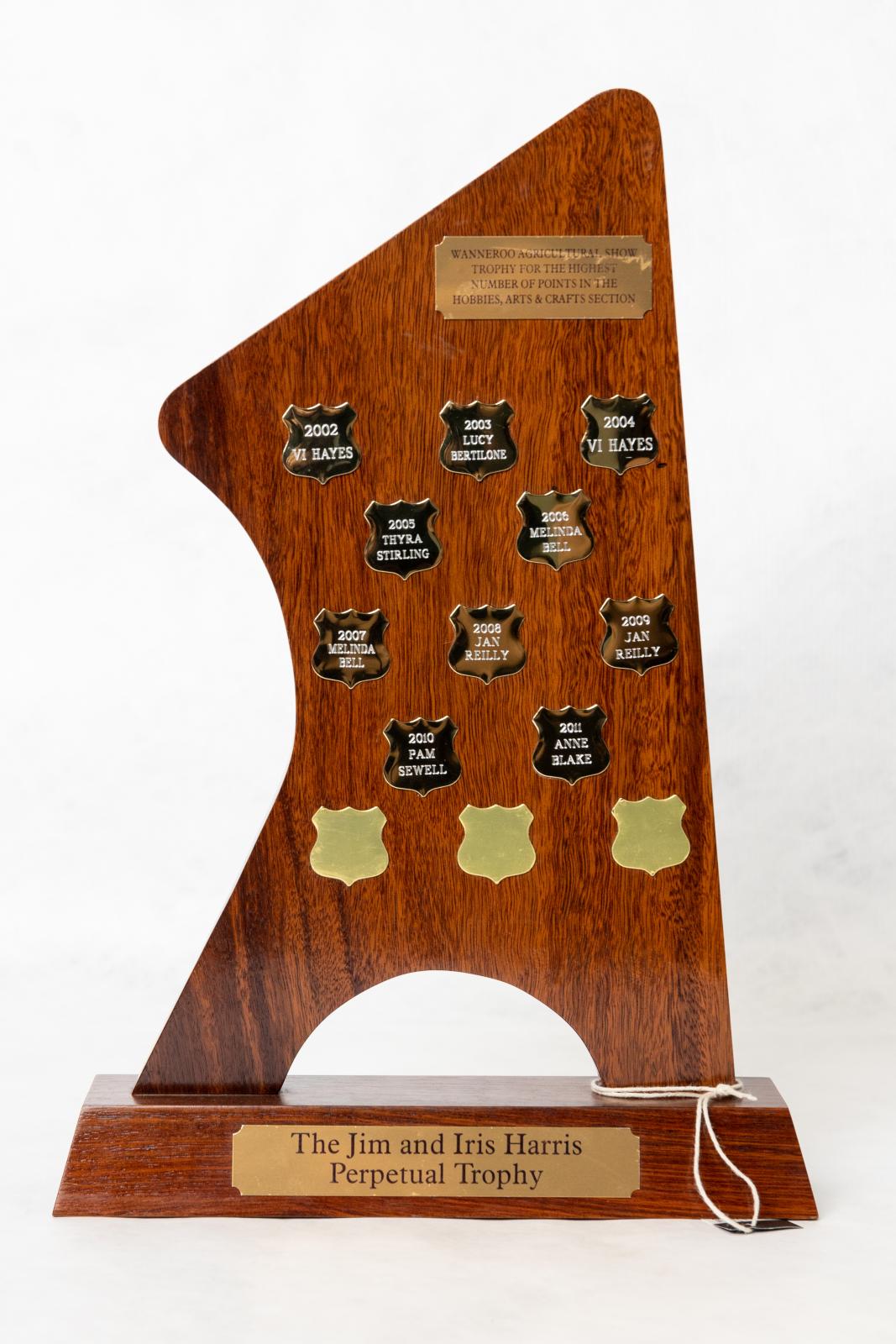Wanneroo Agricultural Society trophy, a point shaped wood board on a wooden base. The trophy has 13 small metal shields attached to the front of the trophy and a metal plaque at the top which explains what the trophy is for. There is a metal plaque on the base with the name of the trophy and a plaque on the back which explains the shape of the trophy. 