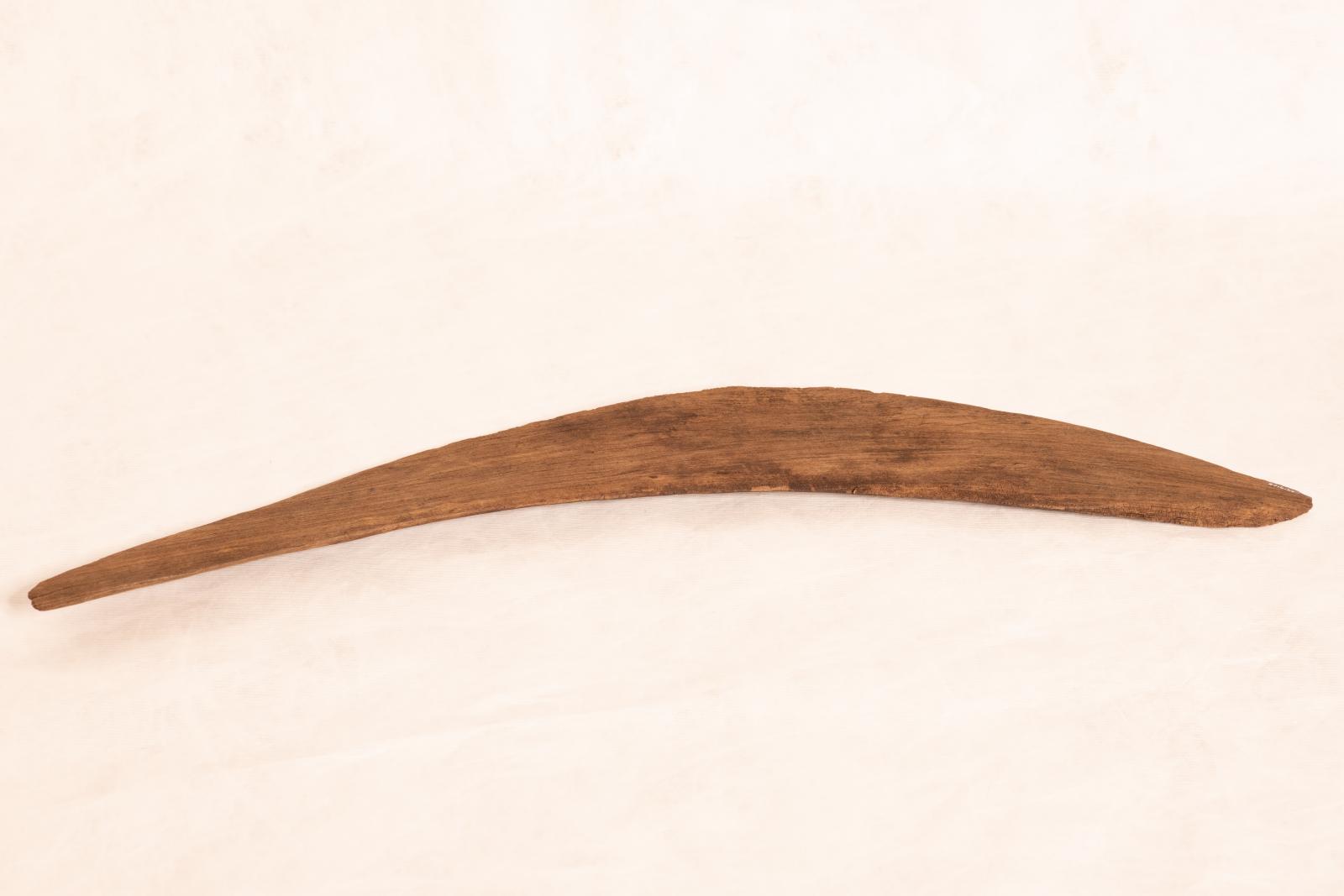 Average sized boomerang, estimated to be 70-100 years old. Slightly curved and thin at one end and thicker at the other. The wood is cracked in multiple areas and on one side the wood has darker patches in areas.