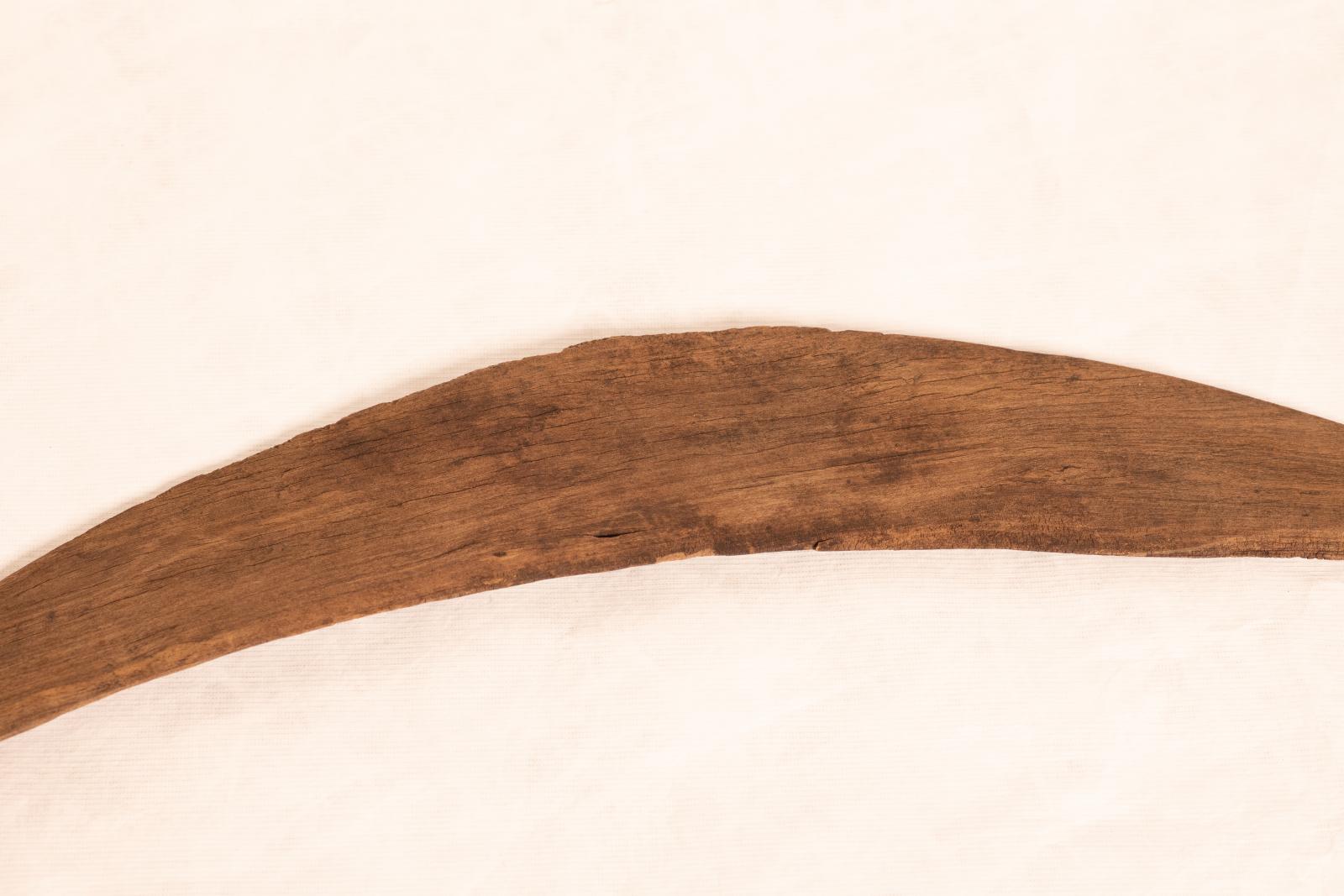 Average sized boomerang, estimated to be 70-100 years old. Slightly curved and thin at one end and thicker at the other. The wood is cracked in multiple areas and on one side the wood has darker patches in areas.