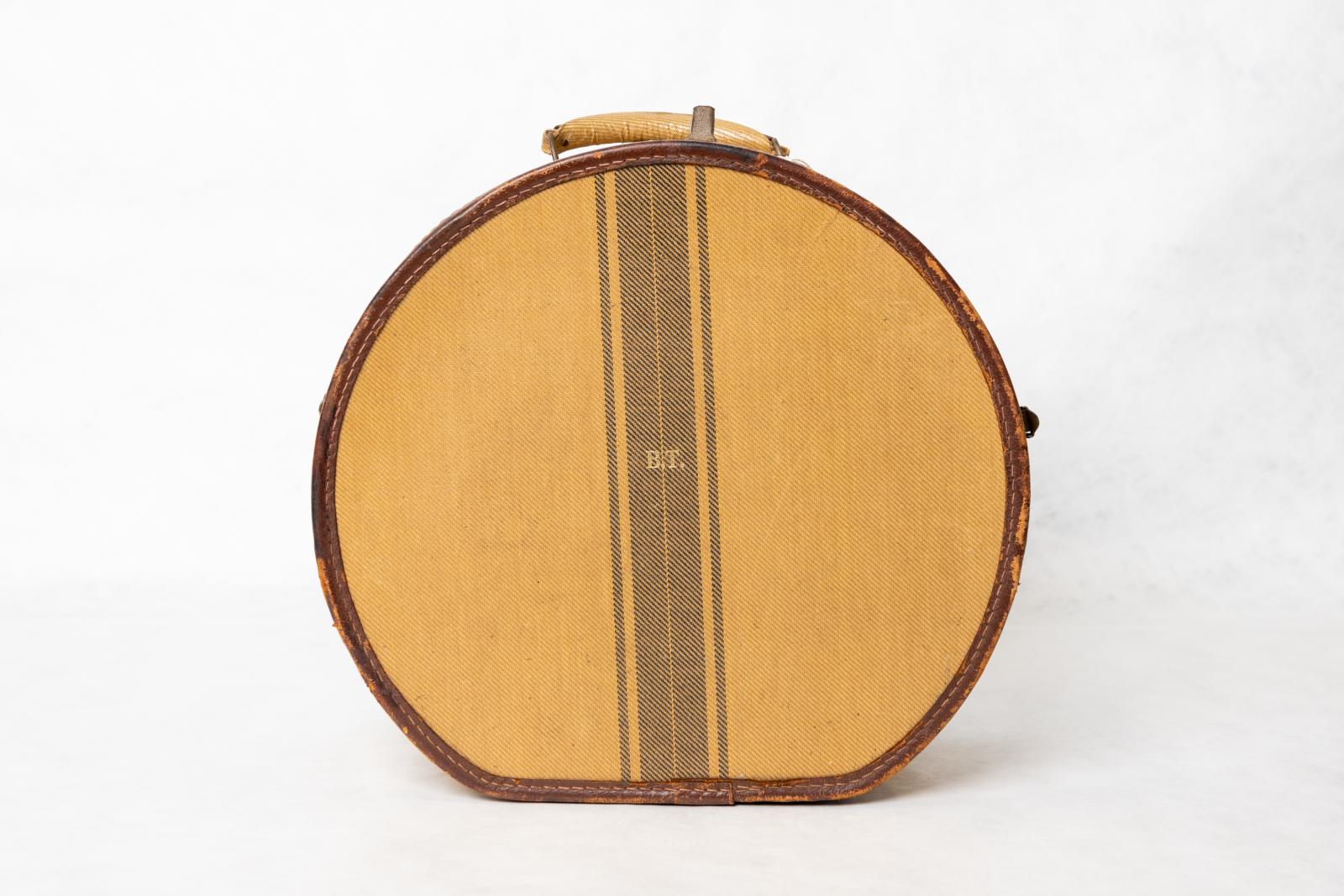 A round sand coloured luggage box with brown edging. There are three brown stripes down the centre of the box, a thick stripe in the middle and two thin stripes on each side. There is a sand coloured handle at the top.