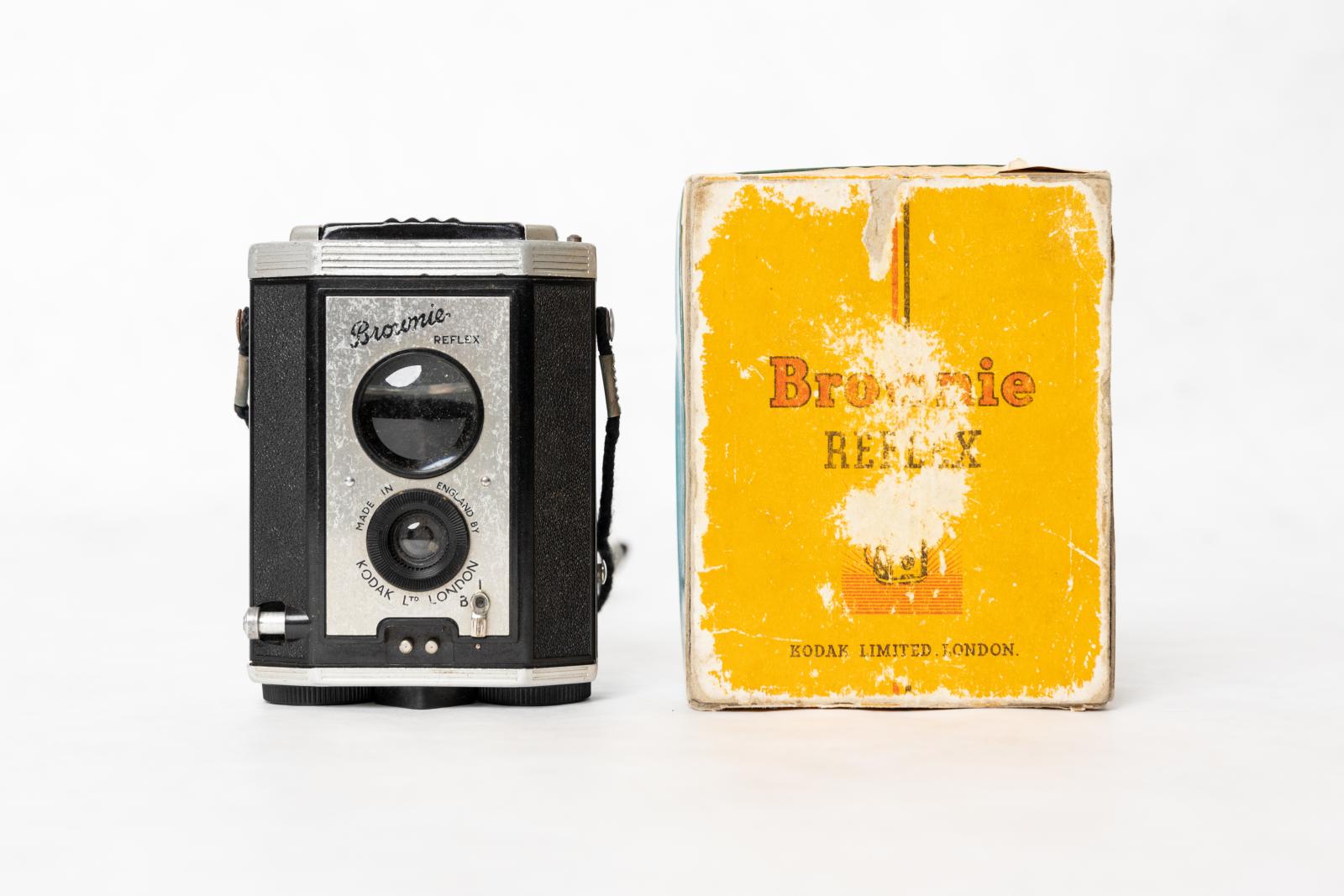 A black and silver rectangular Kodak Brownie camera next to a worn yellow camera box.