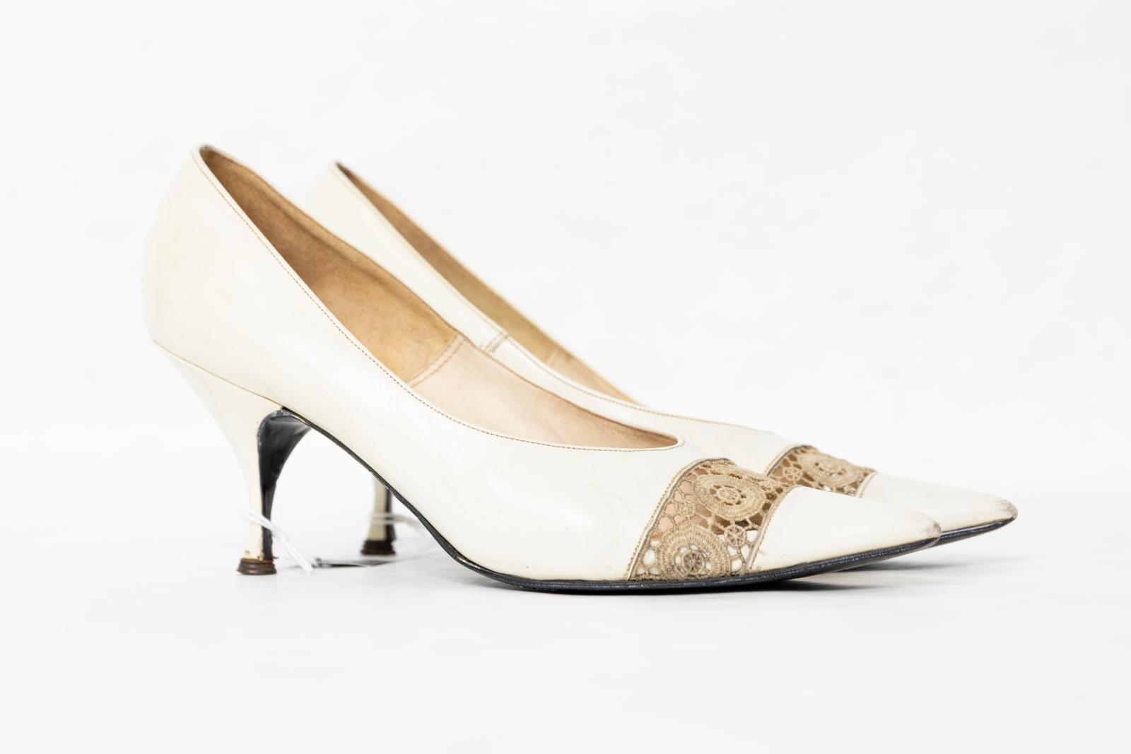 A pair of ladies 'Fern Whispers' shoes sized 6 1/2 with a low stiletto heel. The shoes have a white leather exterior with leather and latex insole, and a black resin sole with a rusted metal tip at the end of each heel. Shoes have pointed toes with a cut-out band over the toe box, which is covered by cream-coloured lace with a circular pattern.