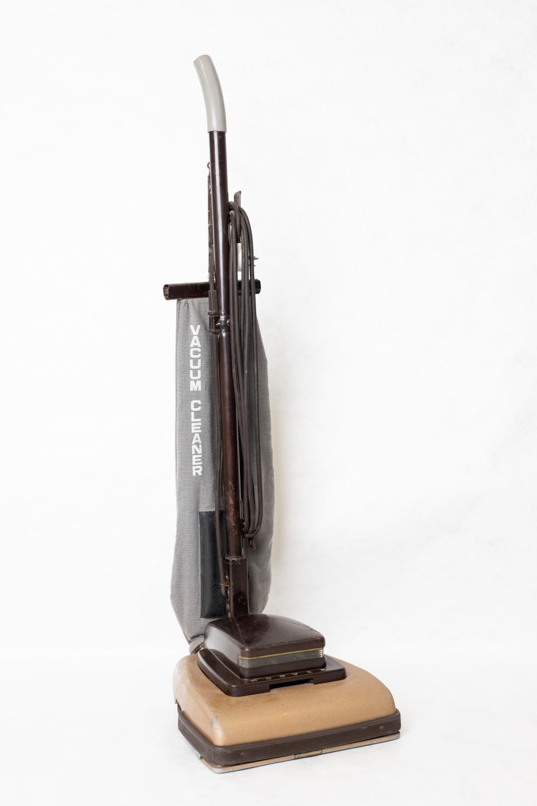 Upright free-standing vacuum. The base of the vacuum is a light brown colour with a dark brown trim around the edge of the bottom, there is a smaller dark brown, square compartment on top of the vacuum base which connects to a long brown handle. The top of the handle has a white plastic end, and the vacuum's power cord is wound around two hooks attached to the handle. Attached to the base of the vacuum is a blue patterned bag to collect the dirt.