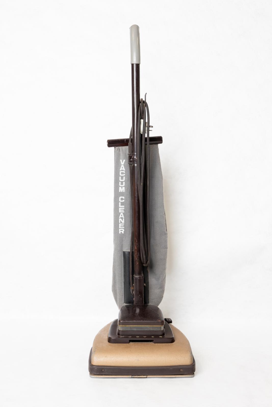 Upright free-standing vacuum. The base of the vacuum is a light brown colour with a dark brown trim around the edge of the bottom, there is a smaller dark brown, square compartment on top of the vacuum base which connects to a long brown handle. The top of the handle has a white plastic end, and the vacuum's power cord is wound around two hooks attached to the handle. Attached to the base of the vacuum is a blue patterned bag to collect the dirt.