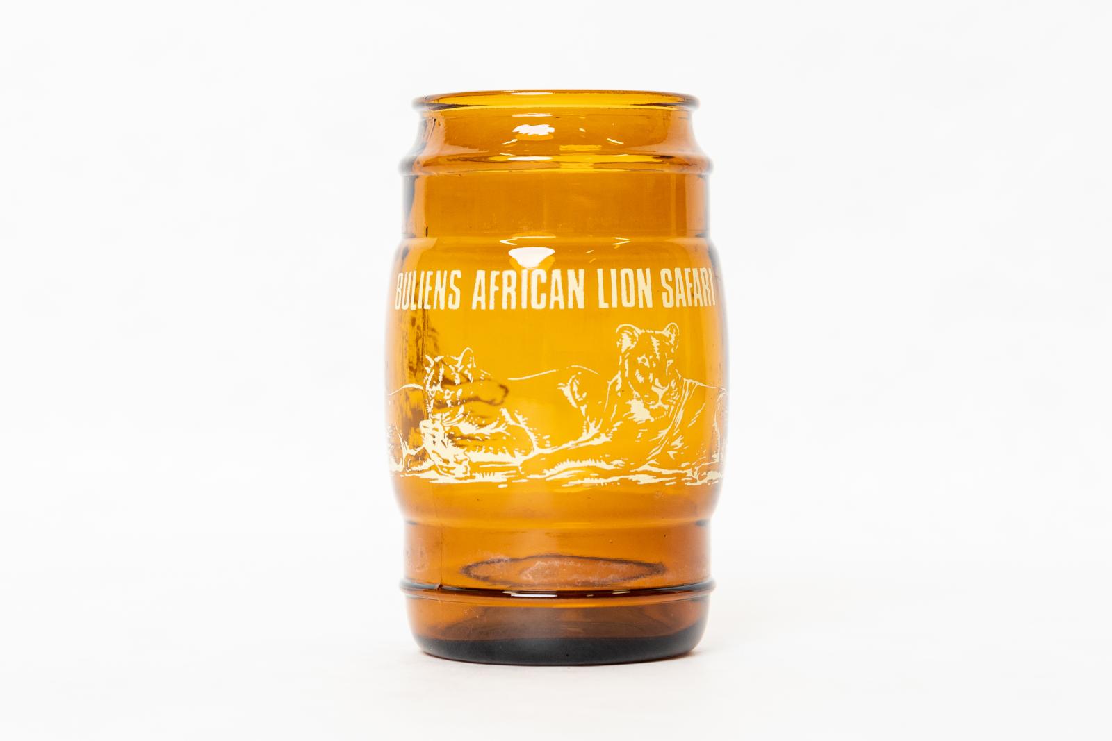 An amber coloured glass with 'BULLENS AFRICAN LION SAFARI' written across the top in white, there is also an outline of lions in white wrapped around the glass. 