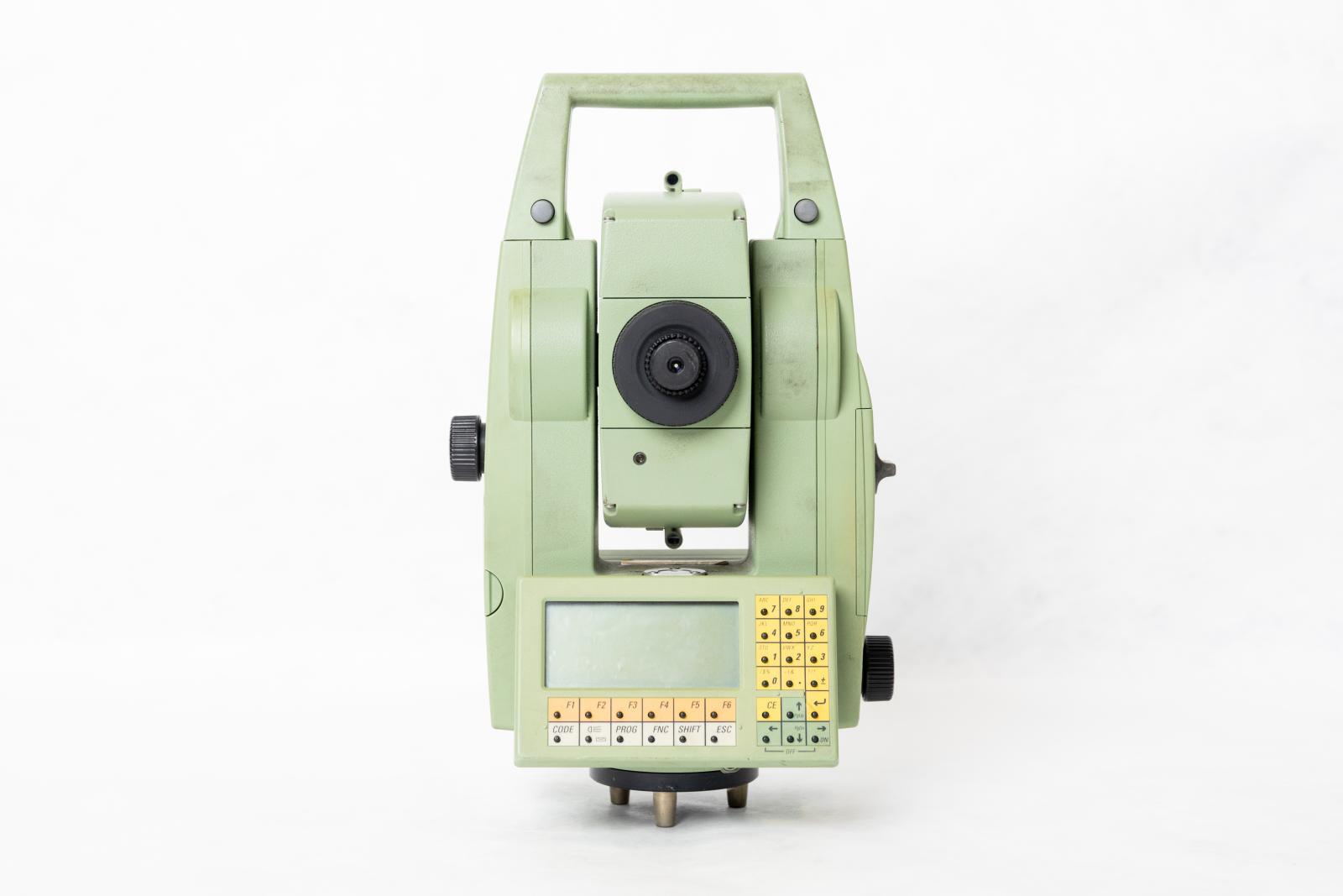 A light green coloured motorized total station with automatic target recognition used for surveying which sits on top of three small metal legs attached to the bottom with a black circle. There is a small screen on both sides of the device at the bottom with a number keypad with arrows on the left and program buttons on the bottom. The device is shaped like a 'U' with a handle on the top. In the centre of the 'U' is a moveable viewing lens. There are also two, one at the b knobs on either side of the device