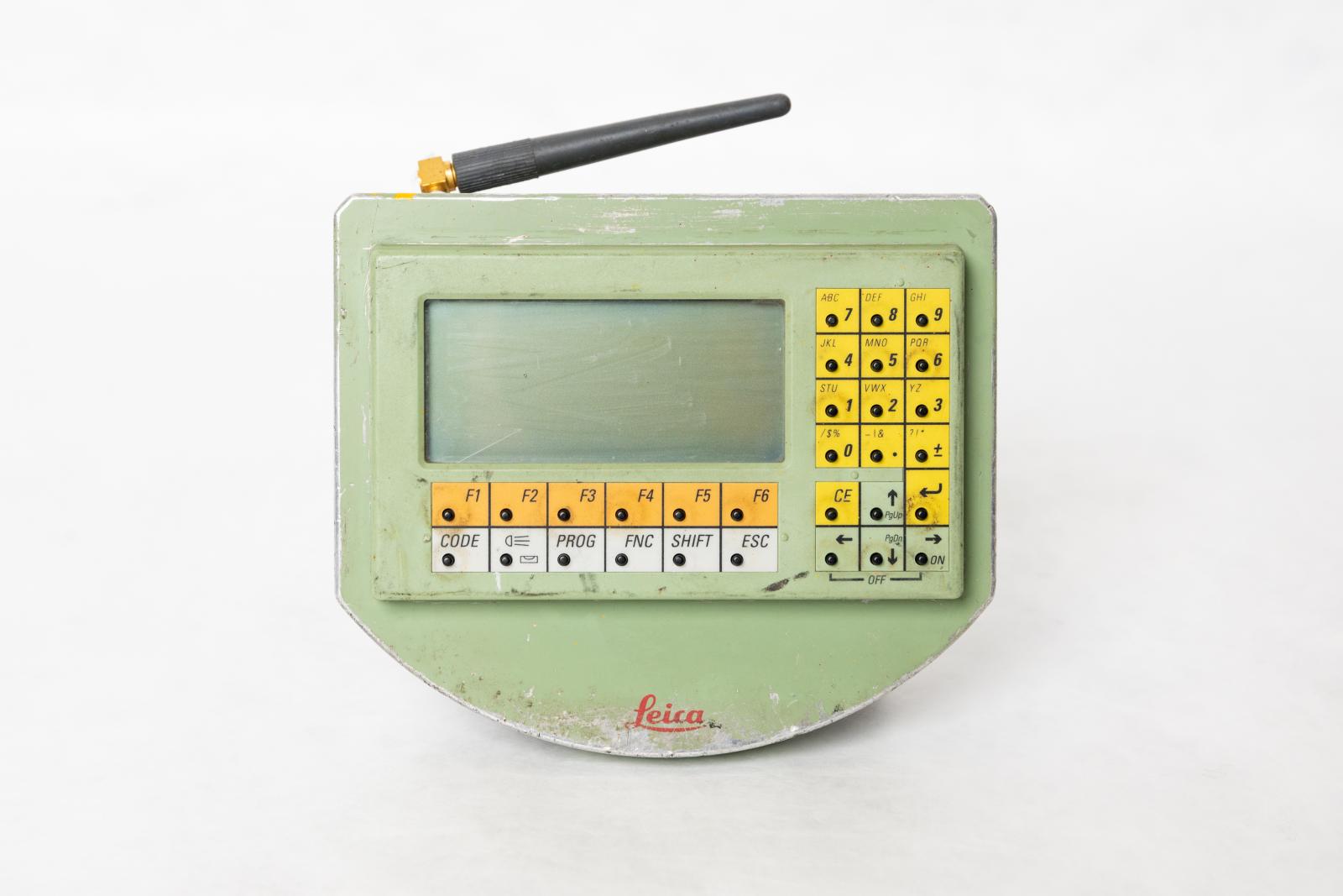 A handheld, light green remote control system with a small screen and program buttons at the bottom of the screen, the top row of buttons is orange and the bottom row is white. On the right hand side of the screen is a number keypad with yellow buttons and below the yellow buttons are arrow buttons in green. Attached to the top of the device is a moveable antennae. 