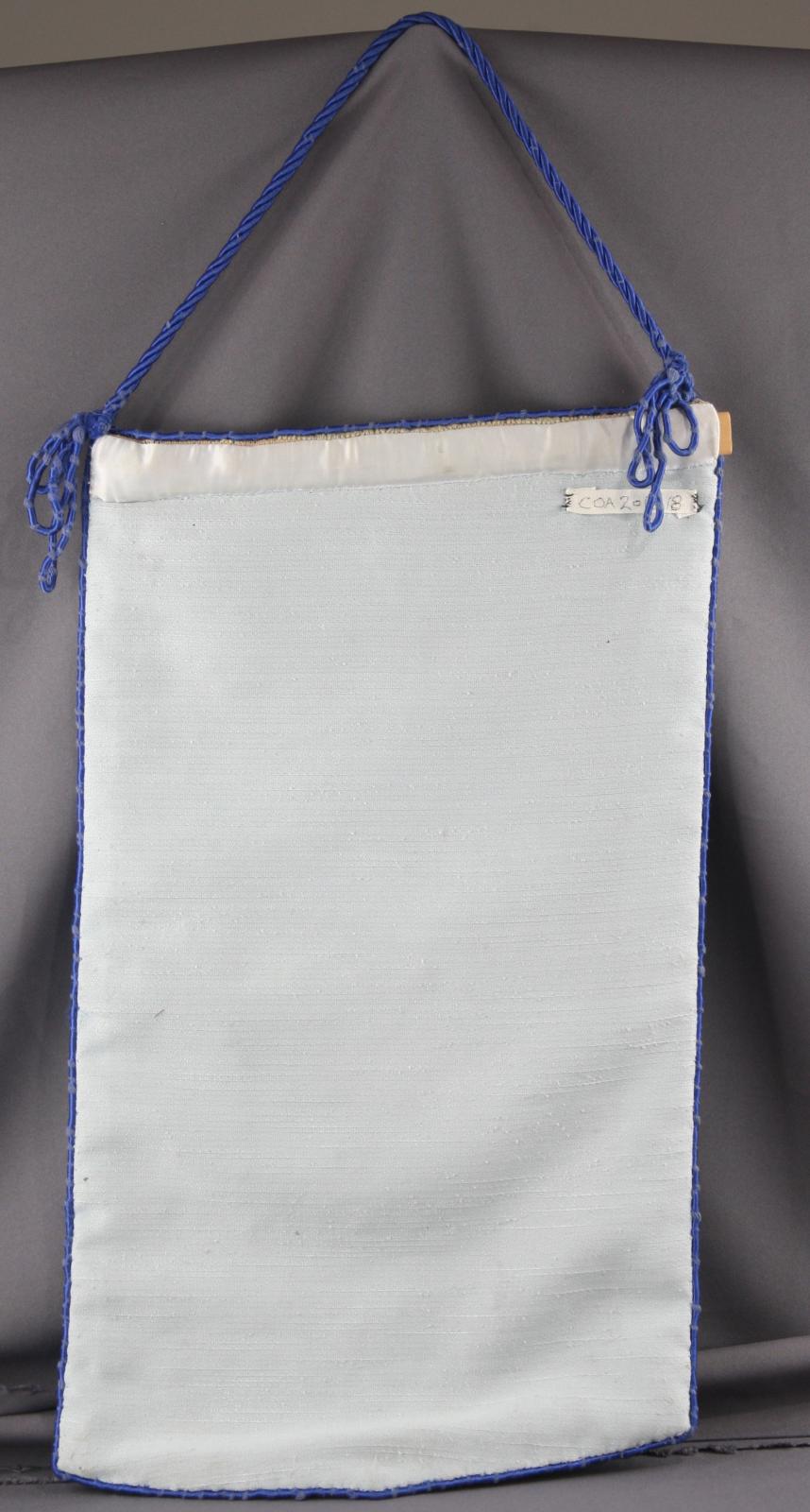 Reverse side of tapestry pennant. The material is light blue colour with dark blue edging around the outside. The pennant is hung by a dark blue cord.