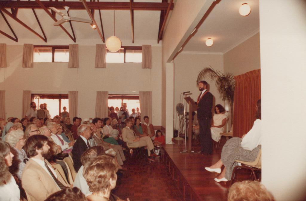 people in a large hall listening to a man speaking from a stage