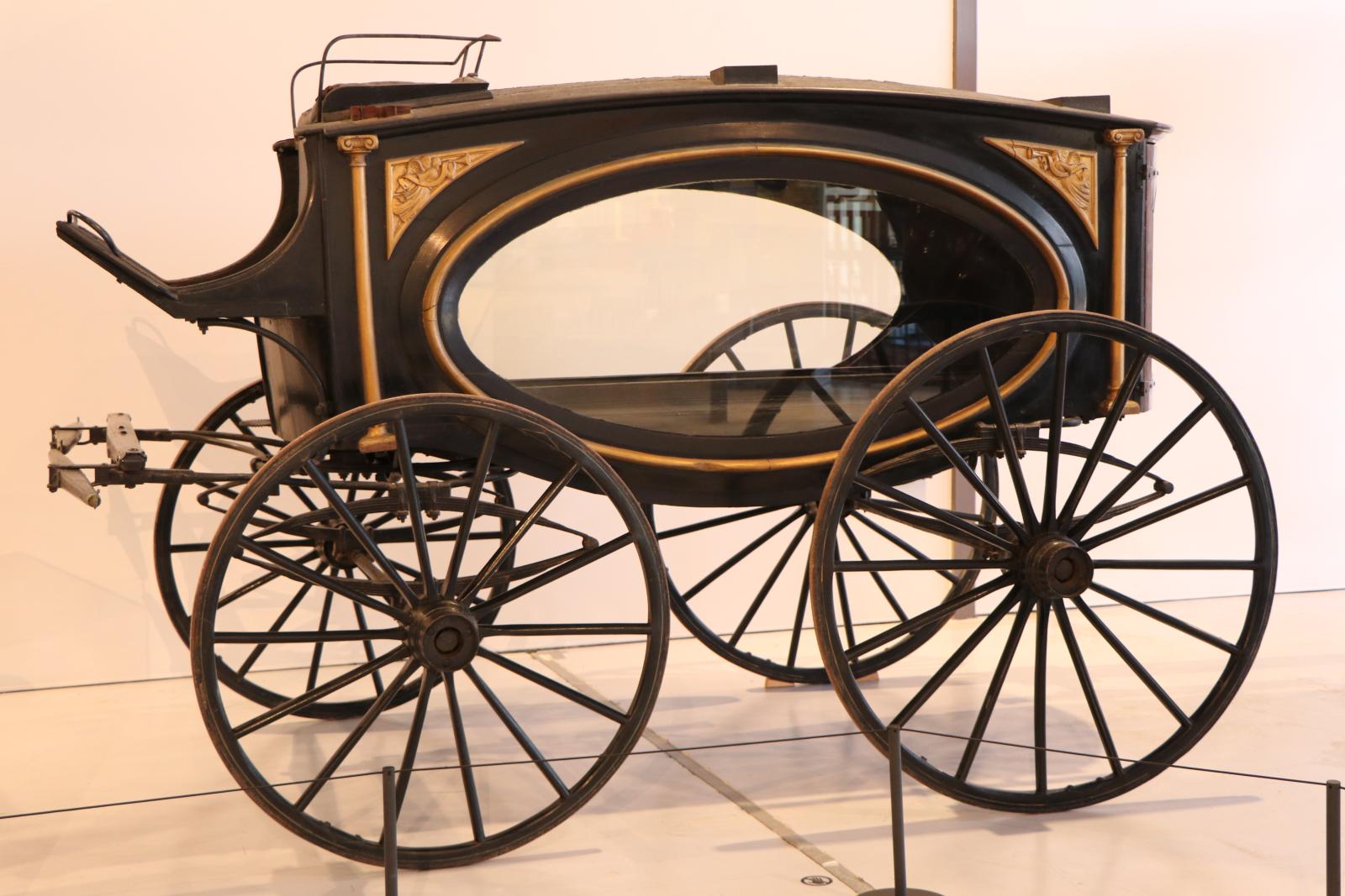 Rectangular black horse-drawn hearse with four wheels. Body painted with gold trimming. Carved golden angels blowing trumpets inset into each corner of body. Two large glass oval windows on both sides of vehicle.