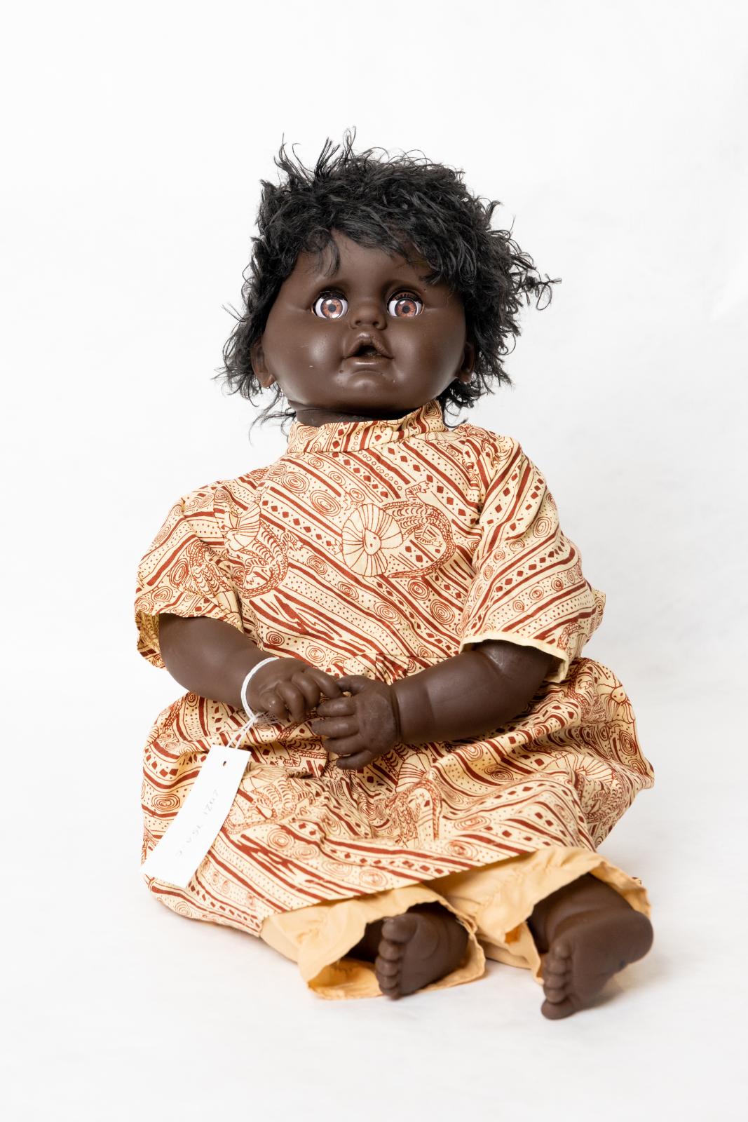 Aboriginal infant girl doll with short black hair. She is wearing a pastel yellow dress with ochre frill neck lizard motif and pastel yellow trousers with ankle frill.