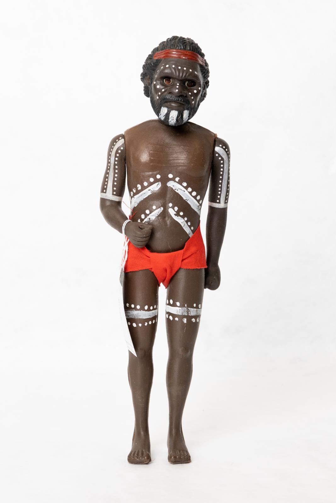 Painted Aboriginal adult male doll wearing a red loin cloth. There is a pattern painted on the dolls torso, arms, legs and face. The doll is wearing a red loin cloth and is wearing a red headband. 