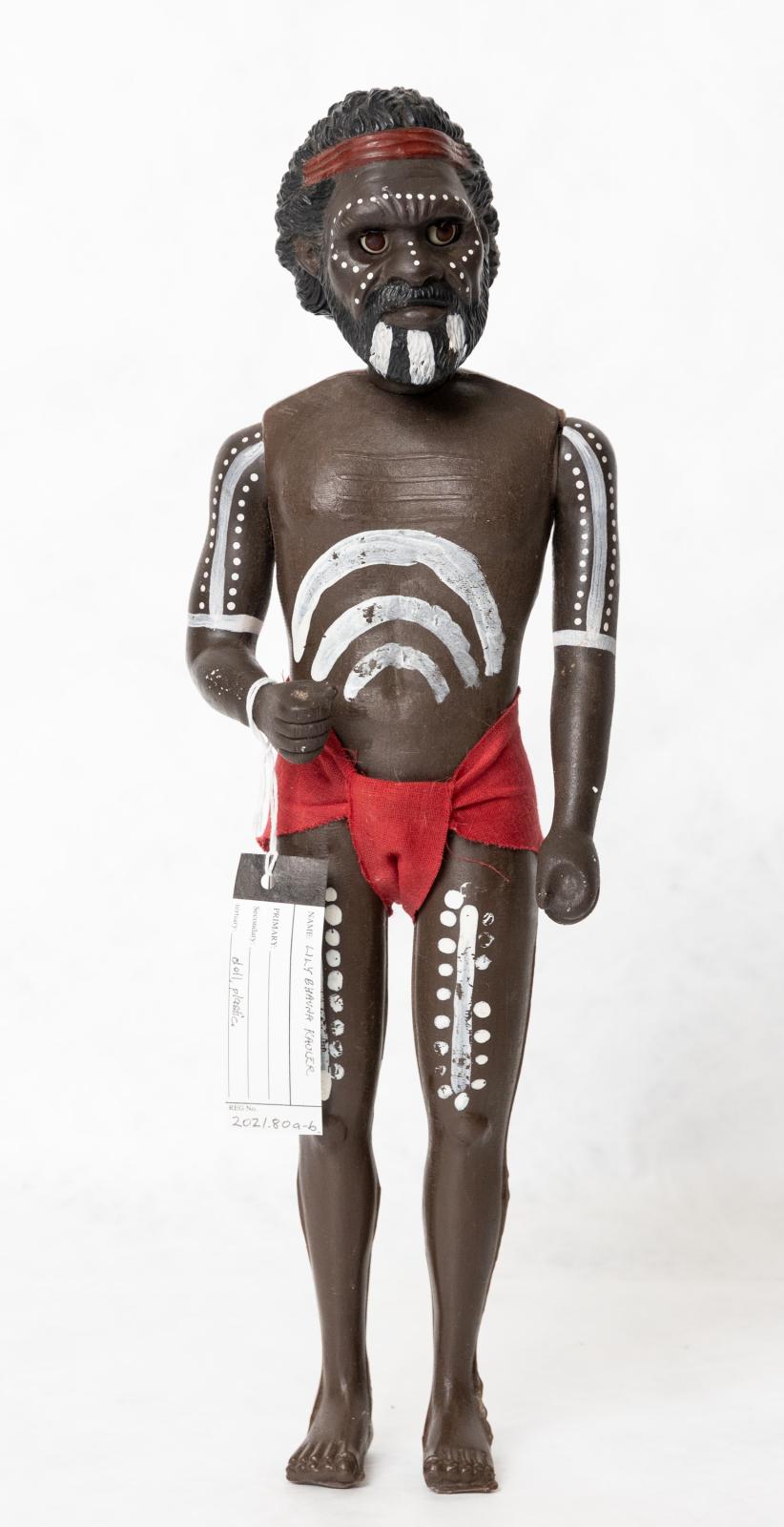 Painted Aboriginal adult male doll wearing a red loin cloth. There is a pattern painted on the dolls torso, arms, legs and face. The doll is wearing a red loin cloth and is wearing a red headband.