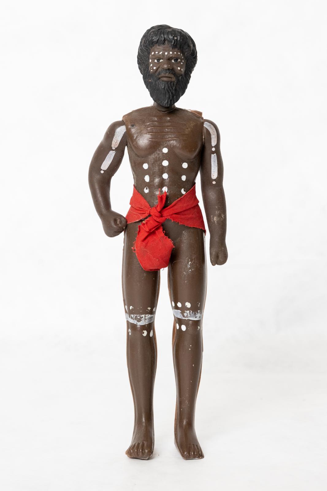 Painted Aboriginal adult male doll wearing a red loin cloth. There is a pattern painted on the dolls torso, arms, legs and face. The doll is wearing a red loin cloth and is wearing a red headband.