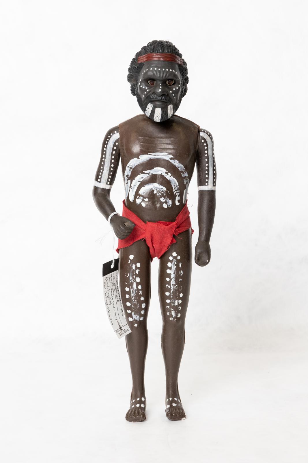 Painted Aboriginal adult male doll wearing a red loin cloth. There is a pattern painted on the dolls torso, arms, legs and face. The doll is wearing a red loin cloth and is wearing a red headband. 