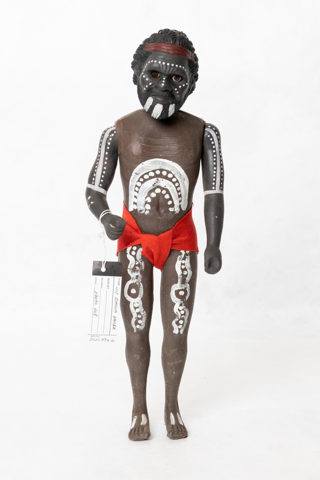 Painted Aboriginal adult male doll wearing a red loin cloth. There is a pattern painted on the dolls torso, arms, legs and face. The doll is wearing a red loin cloth and is wearing a red headband. 