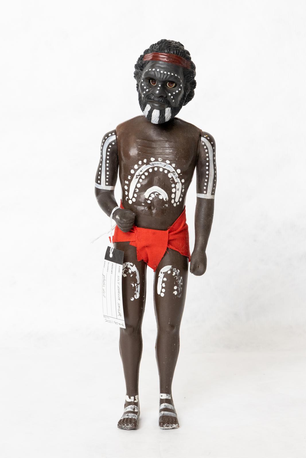 Painted Aboriginal adult male doll wearing a red loin cloth. There is a pattern painted on the dolls torso, arms, legs and face. The doll is wearing a red loin cloth and is wearing a red headband.