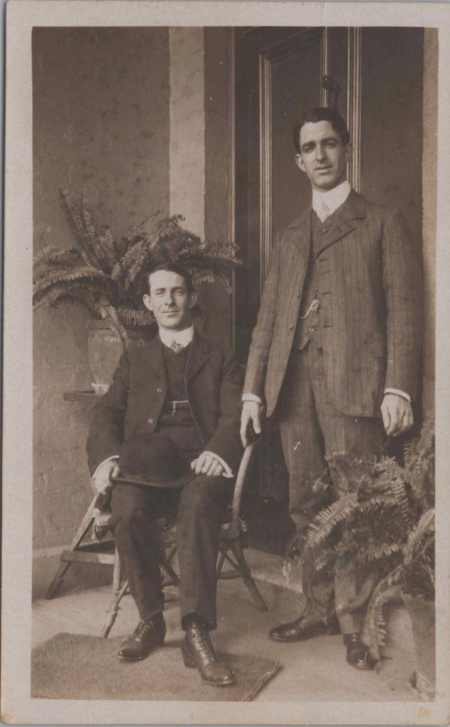 Two gentlemen posing for the camera. One is sitting and one is standing
