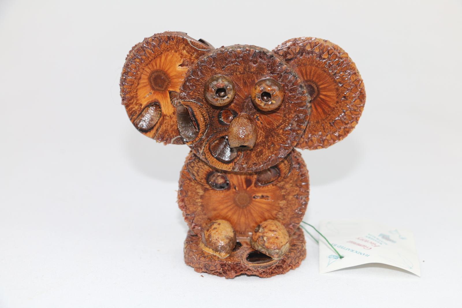 Nut styled Koala glued together with cross-cut sections of banksia in shape of a Koala with seed pods for nose and eyes and feet. A gum nut is located at back of figurine. The object is clear-varnished. Label attached to figurine with cord.