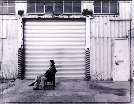 Black and white photograph print of Eveline Kotai sitting on a chair in front of a roller shutter door 